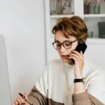 Concentrated short haired female entrepreneur in eyeglasses having discussion via smartphone in modern office