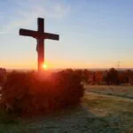 a cross on a hill with the sun setting in the background